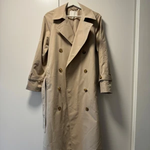Beige trenchcoat med bälte och knappar - Stilren beige trenchcoat med dubbelknäppning och klassisk krage. Jackan har långa ärmar, bälte i midjan och knappar framtill. Perfekt för en trendig och tidlös look. Pris kan diskuteras vid snabb affär