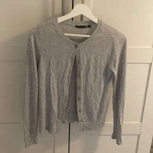 Ljusgrå kofta från 157 - Ljusgrå kofta från 157 i mjuk bomullsblandning. Klassisk rund halsringning och knäppning framtill med små knappar. Långärmad modell med ribbade muddar vid ärmslut och nederkant. Perfekt att slänga över en topp.