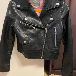 Outwear by Lindex svart jacka, storlek 140 - Svart jacka med dragkedja och tryckknappar. Jackan har två fickor med dragkedja och en fakeficka med tryckknapp. Fodret är rosa. Den har defekter på ärmsluten, se bild 4 och 5.