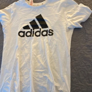Vit t-shirt från Adidas - Säljer en klassisk vit t-shirt från Adidas med deras ikoniska logga i svart på framsidan. T-shirten har en normal passform och korta ärmar, perfekt för en sportig look. Passar bra till både träning och vardag.