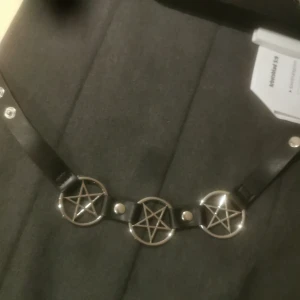 Svart bälte med pentagramdetaljer - Coolt svart bälte med tre pentagramdetaljer i silver. Bältet har tryckknappar för justering och är perfekt för att ge en edgy touch till din outfit. Passar bra till både jeans och klänningar.