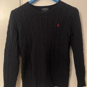 Svart kabelstickad tröja från Ralph Lauren - Säljer en stilren svart kabelstickad tröja från Ralph Lauren med det klassiska röda logotypen på bröstet. Tröjan har långa ärmar och en rund halsringning. Perfekt för en elegant och avslappnad look. 