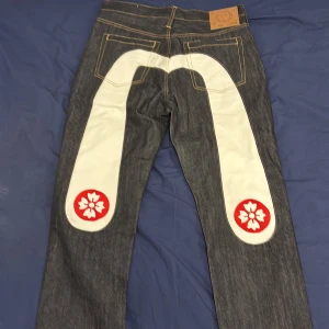 Evisu jeans - Snygga Evisu jeans med en unik vit och röd design på baksidan. De har en klassisk femficksmodell och är tillverkade i mörkblå denim med kontrasterande sömmar. Perfekta för dig som vill sticka ut med en cool och annorlunda stil.