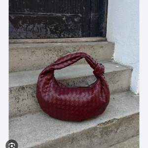 Vinröd flätad handväska - Snygg vinröd handväska med flätat mönster och rundad form. Väskan har ett kort handtag som gör den enkel att bära. Perfekt för att ge en touch av stil till din outfit.