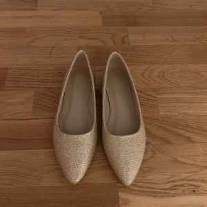 Glittriga guldiga/champagne ballerinaskor - Snygga glittriga ballerinaskor i guld/champagne med spetsig tå. Perfekta för att lägga till lite glamour till din outfit. De har en elegant design och är bekväma att bära. vet inte exakt storleken men ca 38