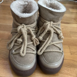 Beige vinterboots från Inuikii - Säljer ett par snygga beige vinterboots från Inuikii. De är tillverkade i mocka med en varm och fluffig insida. Skorna har en robust sula och dekorativa snören som ger en cool look. Perfekta för kalla dagar!