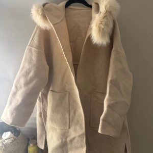 Beige kappa med pälshuva - Säljer en stilren beige kappa med en mysig pälshuva. Perfekt för kyliga dagar. Den har stora fickor framtill och en avslappnad passform. Ett måste för garderoben!