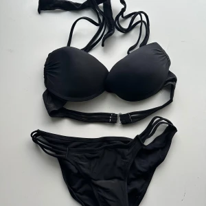 Helt NY Svart bikini med knytband - Snygg svart bikini med knytband i nacken och flera smala band på sidorna av både över- och underdel. Perfekt för en stilren look på stranden eller vid poolen.