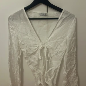 Vit långärmad blus från Design Si - Säljer en elegant vit blus från Design SI med långa ärmar och en v-ringad design. Blusen har en knytning framtill som ger en fin detalj. Perfekt för en stilren look. Storlek xs-s