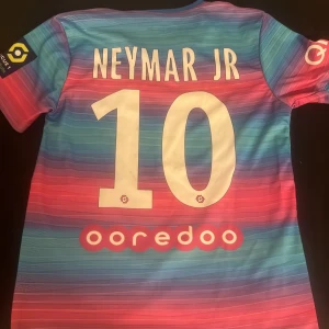 PSG Neymar Jr fotbollströja - Snygg PSG fotbollströja med Neymar Jr och nummer 10 på ryggen. Tröjan har en cool färgkombination av blått och rosa med korta ärmar. Den har PSG:s emblem och sponsorloggor på framsidan. Perfekt för fans av Neymar och PSG!