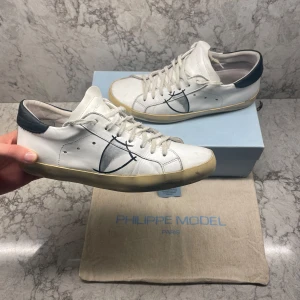 Philippe Model Psrx sneakers - Snygga vita sneakers från Philippe Model med svarta detaljer och snörning. Skorna har en klassisk design med en kontrasterande mörk häl och en bekväm sula. Perfekta för en stilren look. Skicket är som nytt utöver att limmet under skon släppt på vissa ställen som visas på bild. 