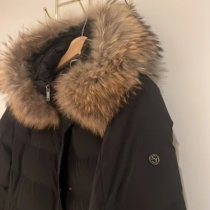 Svart dunjacka med pälsdetalj - Snygg svart dunjacka med en stor, fluffig pälsdetalj runt huvan. Jackan har långa ärmar och stängs med både dragkedja och knappar. Perfekt för kalla dagar när du vill hålla dig varm och stilren. 🖤 äkta päls 