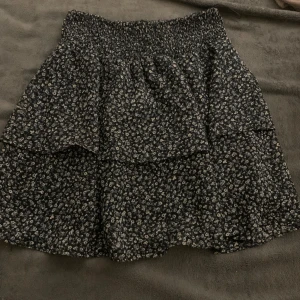 Blommig kjol från Pigalle - Söt blommig kjol från Pigalle i svart och vit. Kjolen har en elastisk midja och volangdetaljer som ger en fin rörelse. Perfekt för en avslappnad stil.