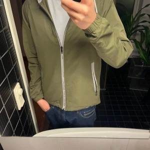 Mörkgrön vindjacka  - Säljer en olivgrön vindjacka med huva och dragkedja framtill. Jackan har långa ärmar och två fickor med dragkedja. Perfekt för blåsiga dagar. 💚 Storlek 48, passar S/M