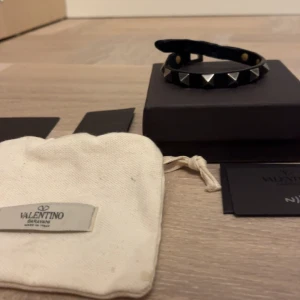 Valentino armband - Snyggt svart armband från Valentino med silverfärgade pyramidnitar. Armbandet har ett stilrent spänne med märkets logotyp. Perfekt för att ge en edgy touch till din outfit.
