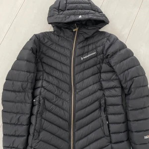 Svart dunjacka från Peak Performance - Snygg svart dunjacka från Peak Performance med huva och dragkedja. Jackan har en quiltad design och är perfekt för kyligare väder. Den har en stilren look med diskreta logotyper på bröstet och ryggen.