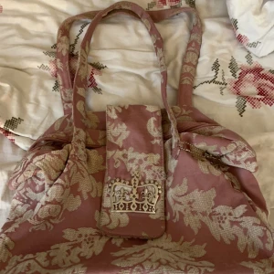 Rosa blommig handväska - Charmig rosa handväska med blommigt mönster och dekorativa detaljer. Väskan har dubbla handtag och en praktisk dragkedja för säker förvaring. Perfekt för att ge en romantisk touch till din outfit.