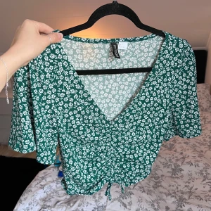 Grön blommig topp - Säljer en söt grön blommig topp från H&M Divided. Toppen har en v-ringad design med korta ärmar och knytning framtill. Perfekt för en somrig look! 🌸💚 Sparsamt använd. Önskar du veta exakta mått? Tveka inte att höra av dig!