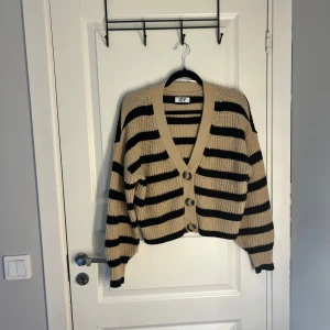 Randig kofta från JDY - Säljer en snygg randig kofta från JDY i beige och svart. Koftan har en v-ringad design med knappar framtill och långa ärmar. Perfekt för en stilren look.