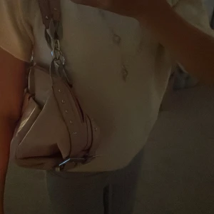 Rosa axelväska med dekorativ detalj - Snygg rosa axelväska med en unik dekorativ detalj framtill. Väskan har en justerbar axelrem med stora metallringar och små nitar som ger en cool look. Perfekt för att ge din outfit en extra touch.