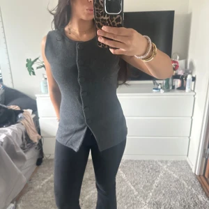 Grå ärmlös topp med knappar - Snygg grå ärmlös topp med knappar framtill. Perfekt för en stilren look. Toppen har en figurnära passform och en djup v-ringning.