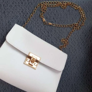 Vit axelväska med guldkedja - Snygg vit axelväska med en elegant guldkedja och ett stilrent lås framtill. Perfekt för att ge din outfit en lyxig touch. Väskan har en kompakt design som gör den lätt att bära med sig.