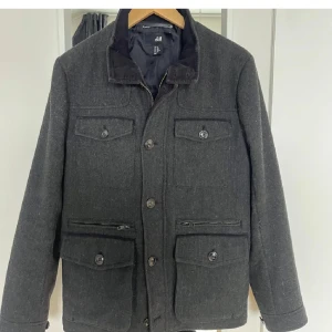 Mörkgrå jacka från H&M - Snygg mörkgrå jacka från H&M med knappar och flera fickor framtill. Jackan har en klassisk krage och långa ärmar, perfekt för kyligare dagar. Den har en stilren design som passar till många olika outfits.
