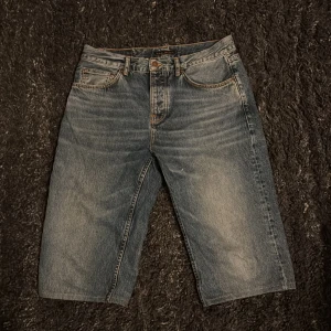 Jeansshorts från Nudie Jeans - Säljer ett par coola jeansshorts från Nudie Jeans. De är i en klassisk blå denimfärg med femficksdesign och knappgylf. Perfekta för en avslappnad stil. Waist är 32 men längden passar nog någon som är 190-200.