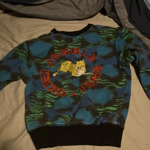 Mönstrad tröja från Kenzo x H&M - Cool tröja från Kenzo x H&M med ett djärvt djungelmönster i blått och grönt. Tröjan har en broderad tiger och texten 'Jungle Kenzo - Paris' i rött på framsidan. Perfekt för dig som vill sticka ut!