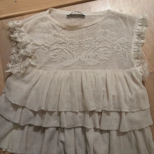 Beige spetsblus med volanger - Charmig beige blus med spetsdetaljer och volanger. Den är ärmlös och har ett vackert blommönster på bröstet. Perfekt för en romantisk look och perfekt till våren och sommaren!💖💖
