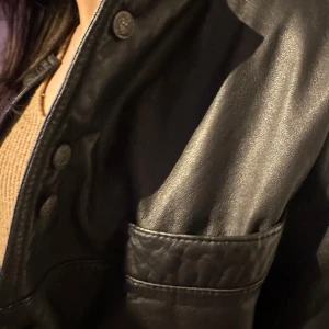 Svart skinnjacka med knappar - Snygg svart skinnjacka med knappar framtill och en cool orange mönstrad insida. Jackan har en stilren design med långa ärmar och två fickor på bröstet. Perfekt för att ge din outfit en edgy touch.