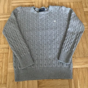 Kabelstickad tröja från Ralph Lauren - Säljer en stilren grå kabelstickad tröja från Ralph Lauren. Tröjan har en broderad logotyp på bröstet och ribbade muddar vid ärmslut och nederkant. Perfekt för kyliga dagar! Storlek: L(14/16) men skulle säga att tröjan passar till en liten xs också