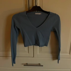 Blå ribbad croppad topp från Stradivarius - Säljer en snygg blå ribbad topp från Stradivarius. Den är croppad med en v-ringad design och har långa ärmar. Perfekt för en stilren look.