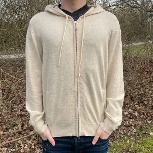 Beige Kashmir zip från Braidbelt UF - Säljer en stilren beige Kashmir hoodie från Braidbelt UF. Tröjan har dragkedja och huva med snörning. Perfekt för en avslappnad look. Passar bra till jeans eller andra casual outfits. Koftan har någon fläck men det går nog enkelt att tvätta bort! Modellen är 180 cm så passar mig som oftast bör M