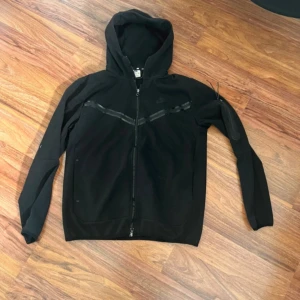 Svart huvtröja från Nike - Säljer en svart huvtröja från Nike med dragkedja och huva. Tröjan har långa ärmar och en praktisk ficka med dragkedja på ärmen. Perfekt för en sportig look eller avslappnade dagar.