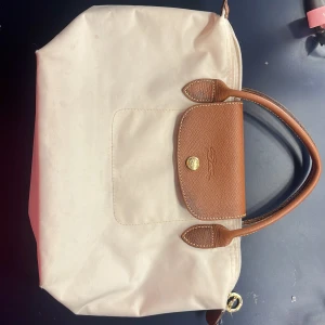 Beige och brun handväska från Longchamp - Säljer en stilren beige handväska från Longchamp med bruna skinnhandtag och detaljer. Väskan har en praktisk dragkedja och en knapp för extra säkerhet. Perfekt för att bära med sig det nödvändigaste med stil. Skriv vid funderingar, kan skicka bättre bilder på dess defekter, mått osv💘