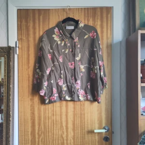 Blommig skjorta från Cappuccini - Snygg, ostruken skjorta från Cappuccini med ett blommigt mönster i rosa och beige på en grön bakgrund. Skjortan har trekvartsärmar och knappar framtill. Perfekt för en avslappnad stil med en touch av elegans. 