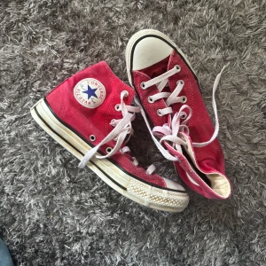 Röda Converse All Star sneakers - Säljer ett par klassiska röda Converse All Star sneakers med vita snören och gummisula. Skorna har den ikoniska loggan på sidan och är perfekta för en avslappnad stil. Några defekter på skosnören och sula då jag målade de med textilfärg från rosa till rött.