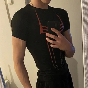 Svart Spimerman Comp. T-shirt - Snygg svart t-shirt med ett coolt spindeltryck i rött och vitt. Perfekt för dig som vill sticka ut med en unik design. T-shirten har en tight passform och korta ärmar, vilket gör den idealisk för träning eller vardagsbruk.