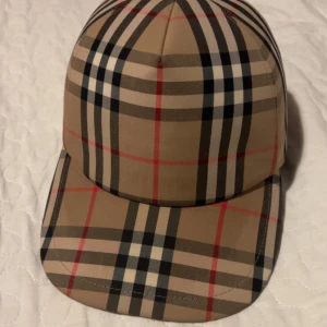  keps från Burberry - Snygg rutig keps från Burberry i klassiskt beige, svart och rött mönster. Kepsen har en justerbar rem baktill för perfekt passform. Perfekt för att ge din outfit en stilren touch.