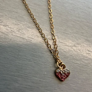 Guldhalsband med hjärthänge - Elegant guldhalsband med ett hjärthänge dekorerat med rosa och vita stenar. Perfekt för att lägga till en gnistrande touch till din outfit. Kedjan är tunn och smidig, vilket gör den bekväm att bära hela dagen.