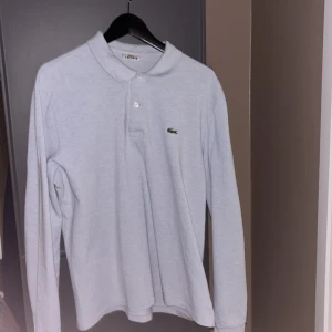 Grå långärmad pikétröja från Lacoste - Säljer en stilren blå/grå långärmad pikétröja från Lacoste med klassisk krage och knappar framtill. Tröjan har det ikoniska krokodilmärket på bröstet. Perfekt för en avslappnad men ändå snygg look. Fråga för fler bilder eller funderingar! Priset är inte skuret i sten!