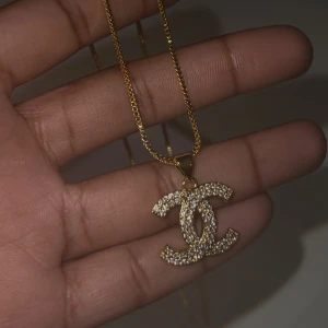 Guldhalsband med glittrig hängsmycke - Elegant guldhalsband med ett glittrigt hängsmycke i form av två sammanflätade C:n. Helt oanvänd, köpt utomlands. Materialet är jag inte säker på.   • Låda medföljer ej, dock paketeras smyckena säkert för leverans