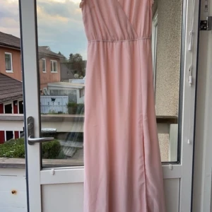 Ljusrosa långklänning - Elegant ljusrosa långklänning med omlott-design och v-ringad hals. Klänningen har korta, fladdriga ärmar som ger en romantisk känsla. Perfekt för sommarens festligheter.