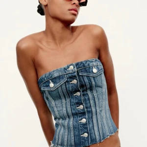 Jeanskorsett Zara - Trendig jeanskorsett i blå denim med knappar framtill och dekorativa fickor. Den är axelbandslös och har en snygg, figurnära passform. Perfekt för att ge en cool touch till din outfit.