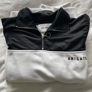 Axel Arigato halfzip - Snygg svart och vit tröja från Axel Arigato med dragkedja och hög krage. Tröjan har en stilren design med märkesnamnet broderat på den vita delen. 