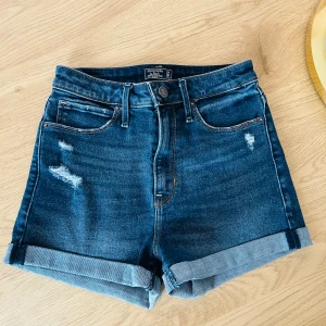 Blå jeansshorts från Abercrombie & Fitch - Snygga blå jeansshorts från Abercrombie & Fitch med hög midja och slitna detaljer. De har en klassisk femficksdesign och uppvikta benslut för en avslappnad look. Perfekta för sommardagar!
