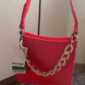 Röd axelväska från HVISK - Snygg röd axelväska från HVISK i återvunnen nylon. Väskan har en cool kedjedetalj och justerbar axelrem. Perfekt för att ge din outfit en färgklick. 🌟Orginal pris 995kr💕