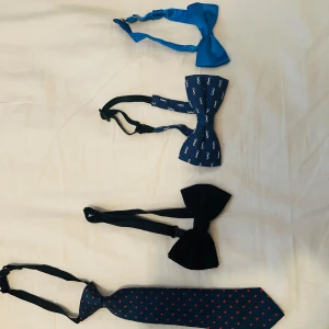 Samling av flugor och slipsar för barn 15kr per styck (60) - Säljer en samling av flugor och slipsar för barn i olika färger och mönster. Inkluderar en blå fluga i satin, en blå fluga med mustaschmönster, en svart fluga och en mörkblå slips med röda prickar. Perfekt för att variera din stil vid olika tillfällen.