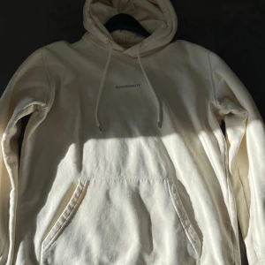 Vit hoodie från NN07 - Säljer en stilren vit hoodie från NN07 med texten 'No Nationality' på bröstet. Hoodien har en klassisk design med känguruficka och justerbar huva.